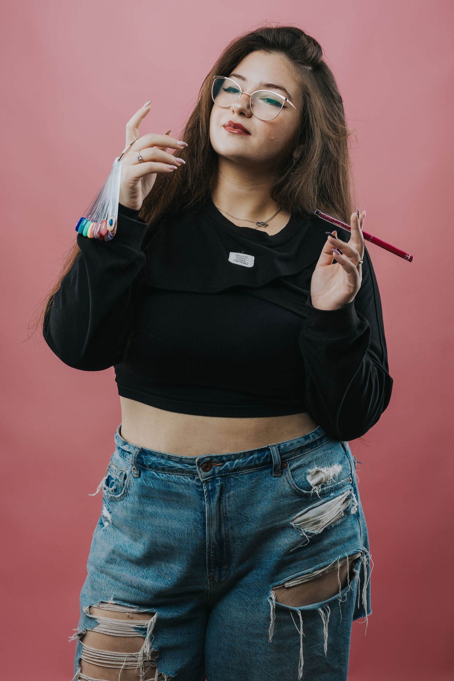 Young woman in crop top and ripped jeans poses confidently against pink background.