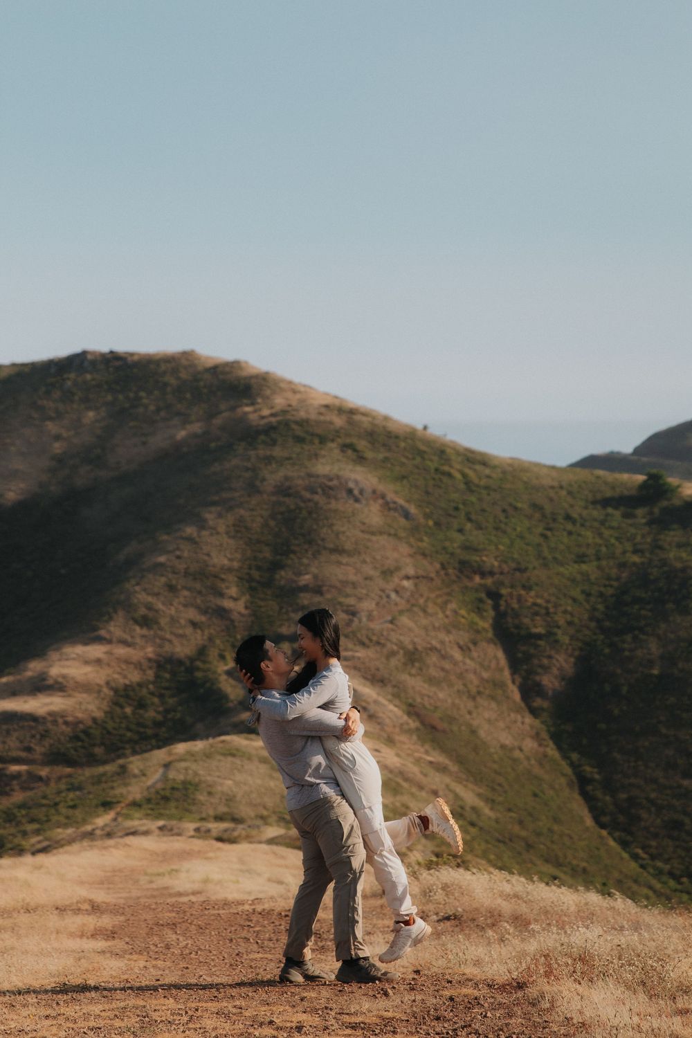 Couple embracing on scenic hilltop trail overlooking rolling coastal mountains on a hazy afternoon.