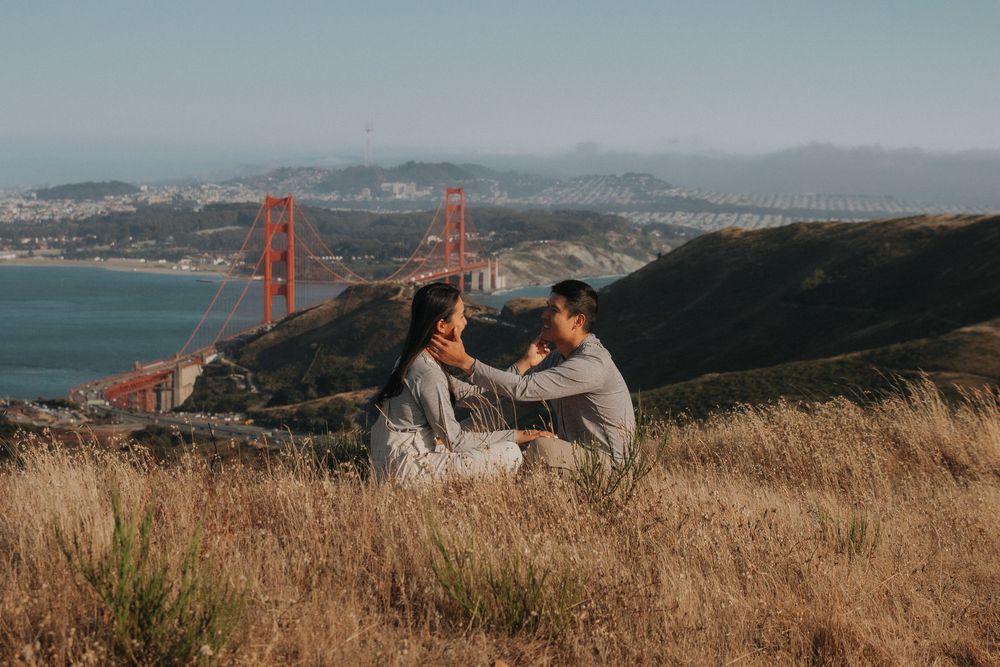 A couple sits in golden grass overlooking the Golden Gate Bridge and San Francisco Bay on a hazy afternoon.