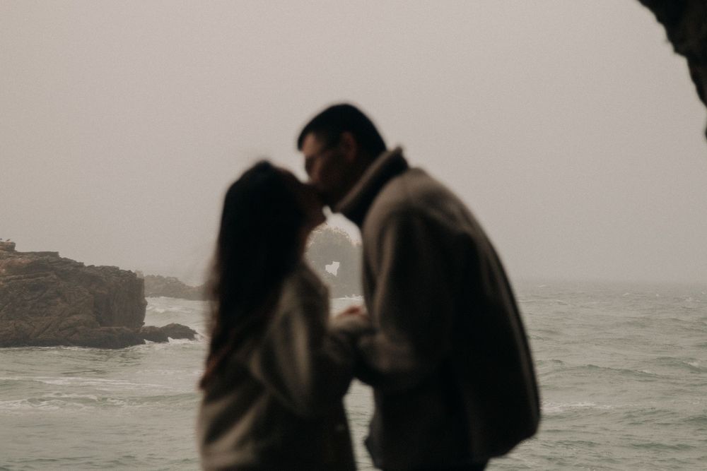 Two silhouetted figures share an intimate moment on a foggy rocky coastline at dusk.