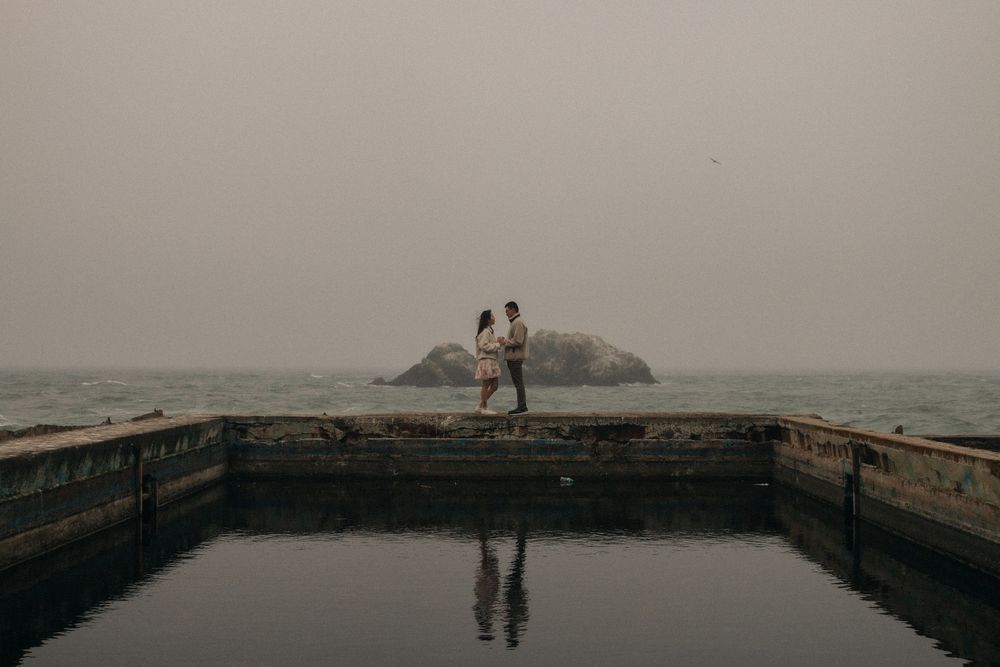 Two silhouettes stand together on the edge of a stone pool overlooking misty sea and rocky landscape at dusk.