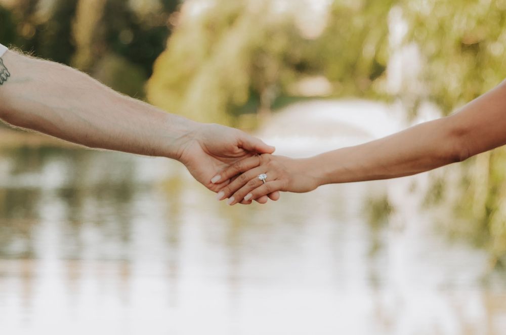 Two hands gently clasped together against a soft blurred nature background with sunlit foliage.