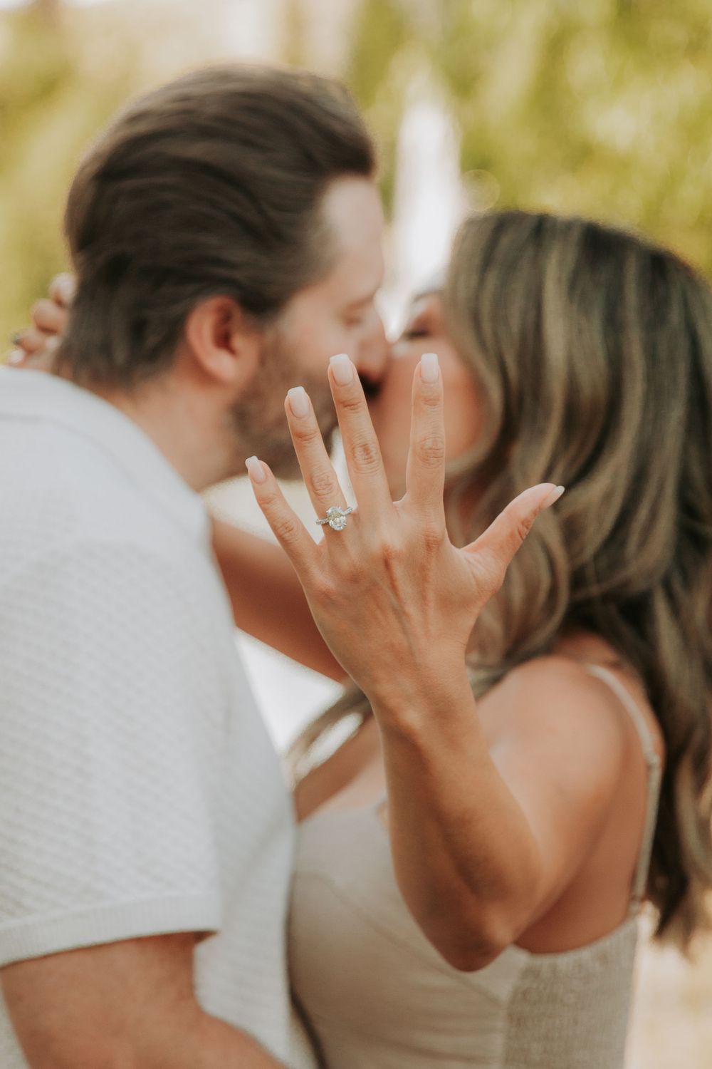 A close-up of an engagement ring on display as a couple shares a romantic moment in the background.