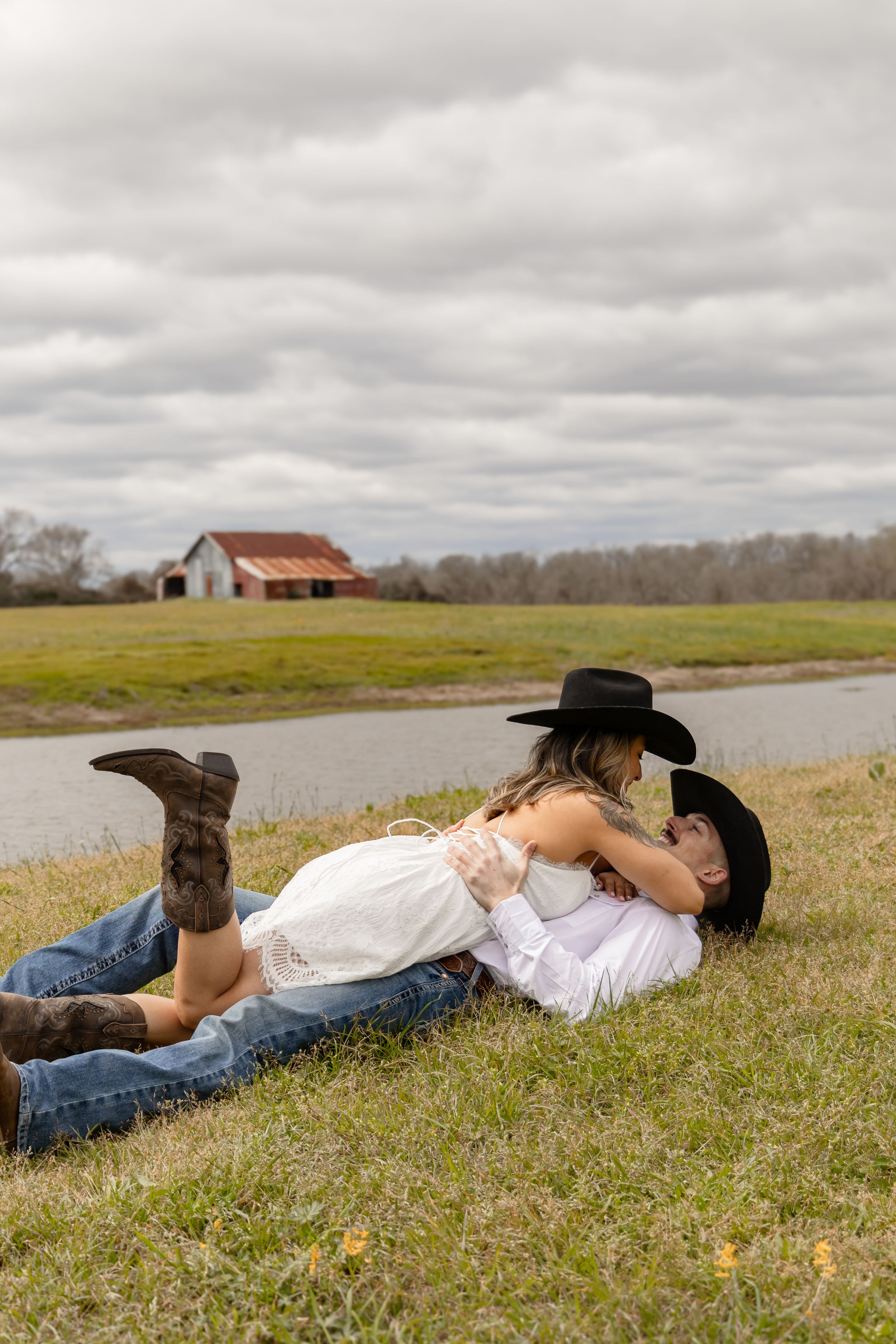 Girl laying on her fiancé in a grassy field with a pond and a barn in the background.