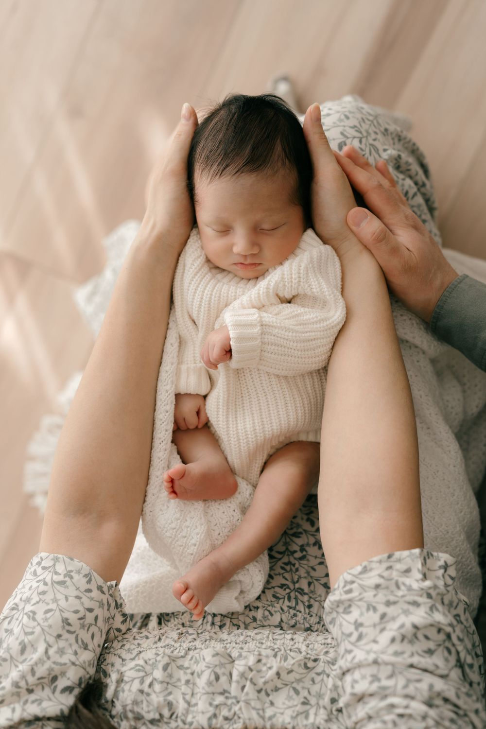 Sleeping newborn baby in white knit onesie resting peacefully between parent's hands on textured blanket.