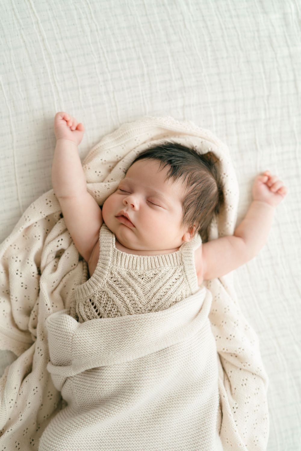 Sleeping infant stretching arms while wrapped in cream knitted blanket on white surface.