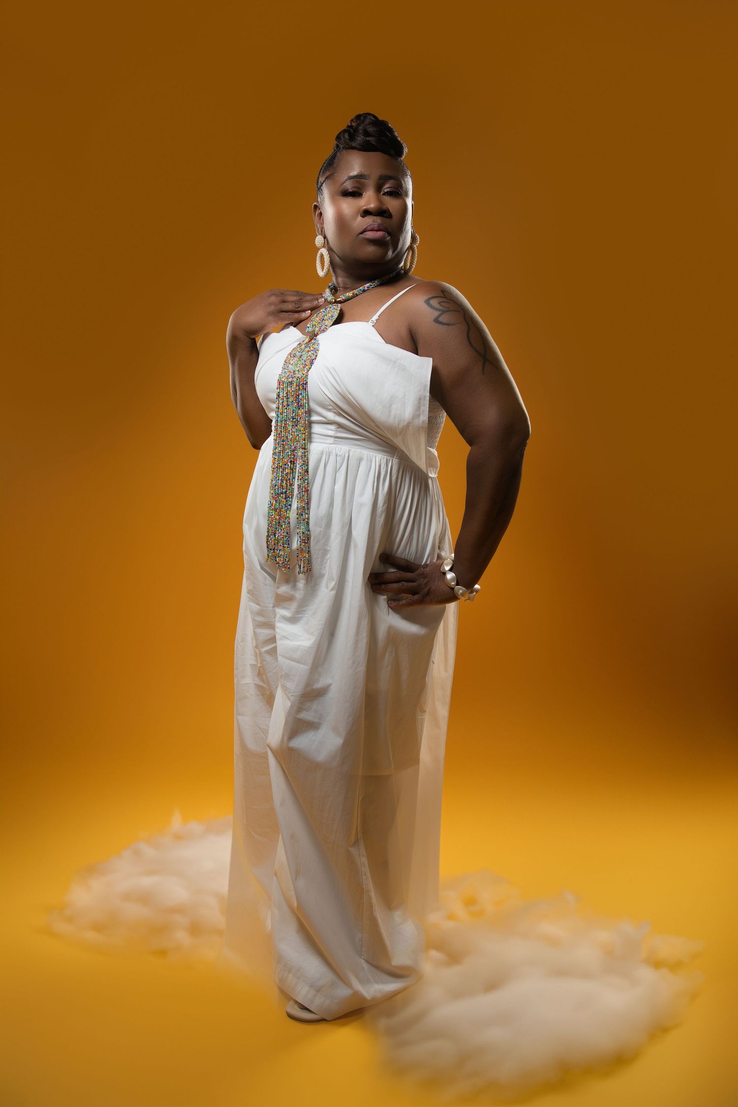 Fashion portrait in flowing white dress with gold accents against orange background.