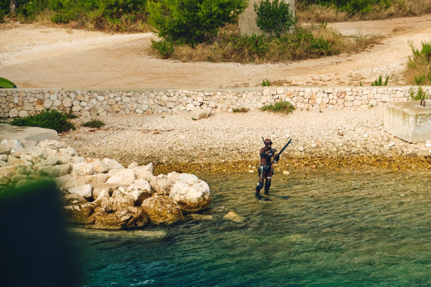 Fisher standing on rocky shoreline casting into crystal clear Mediterranean waters.