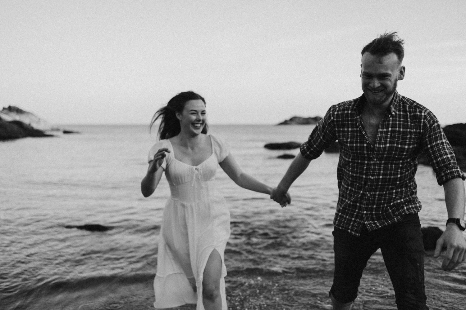 Black and white photo of couple running along rocky shoreline at sunset.