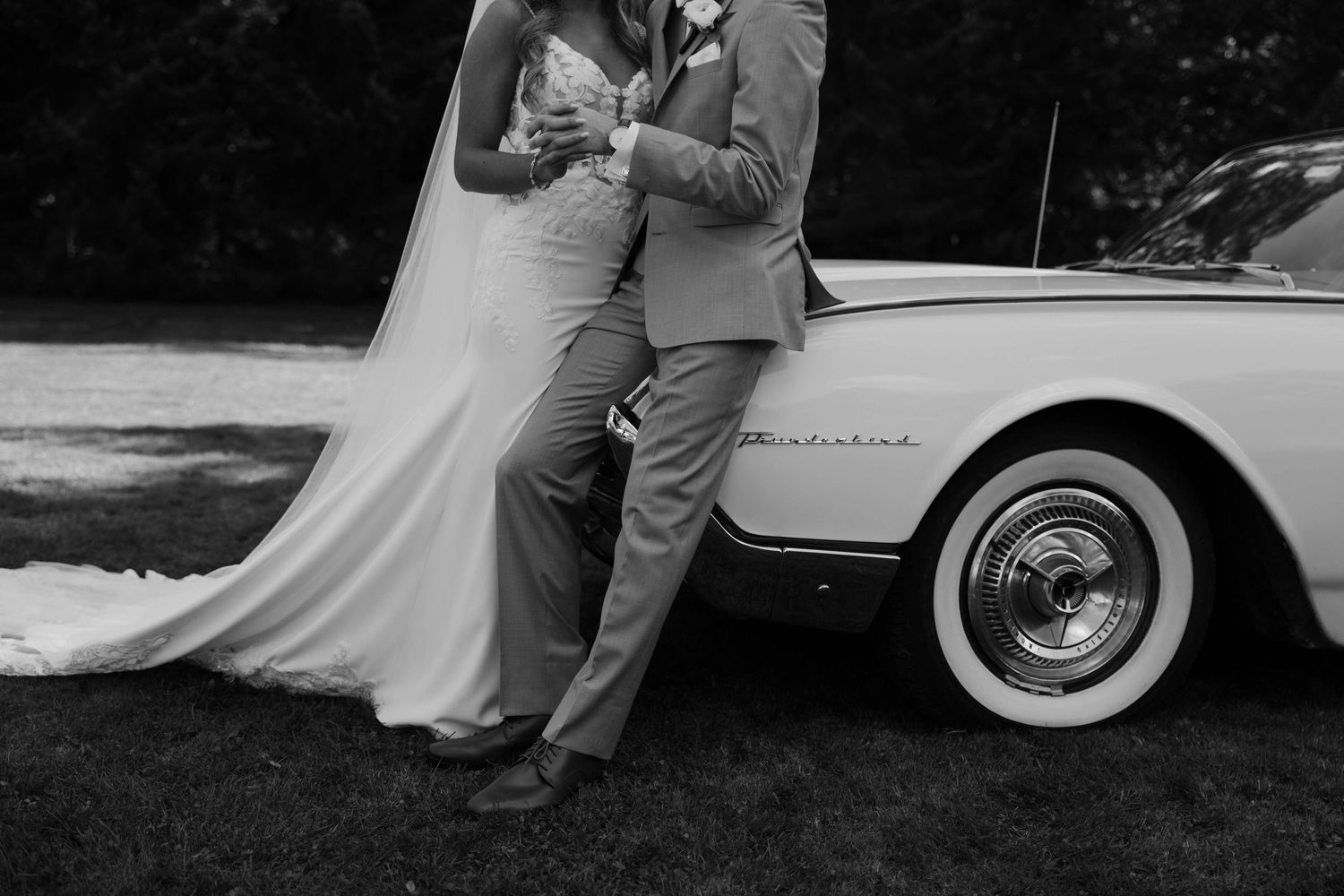 A black and white wedding photo of newlyweds posing beside a vintage car.