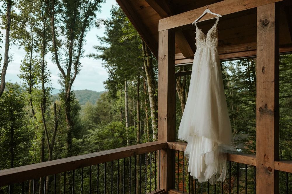 Wedding dresses hang from wooden beams in an outdoor pavilion surrounded by lush forest scenery.