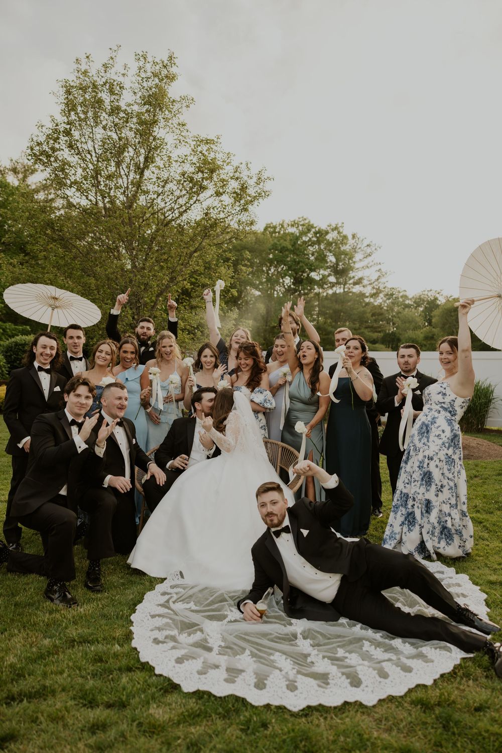Wedding party sits together on grassy lawn during outdoor celebration with bride's white dress spread out beautifully.