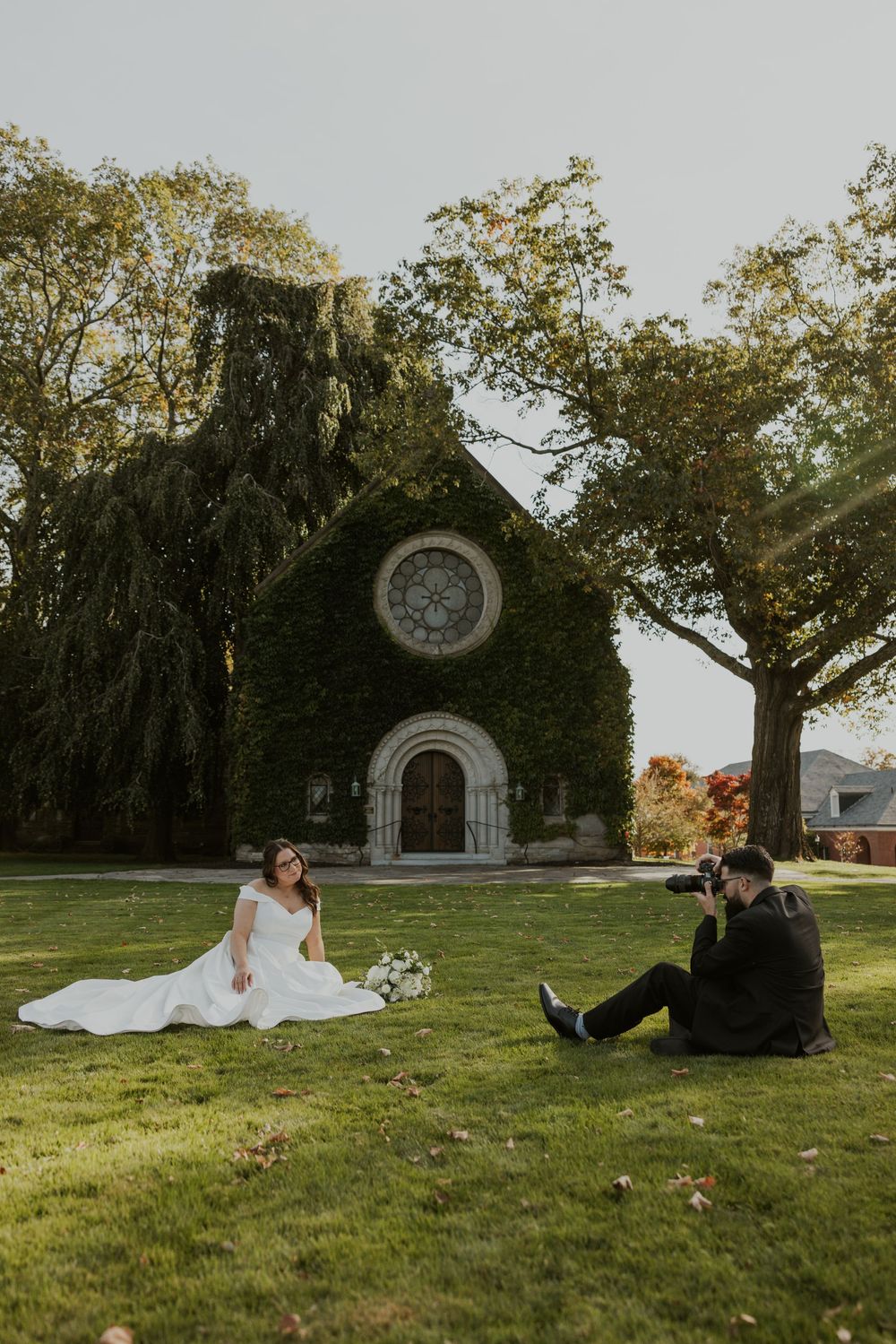 Couple relaxes on grassy lawn in front of stone chapel during outdoor wedding photoshoot.