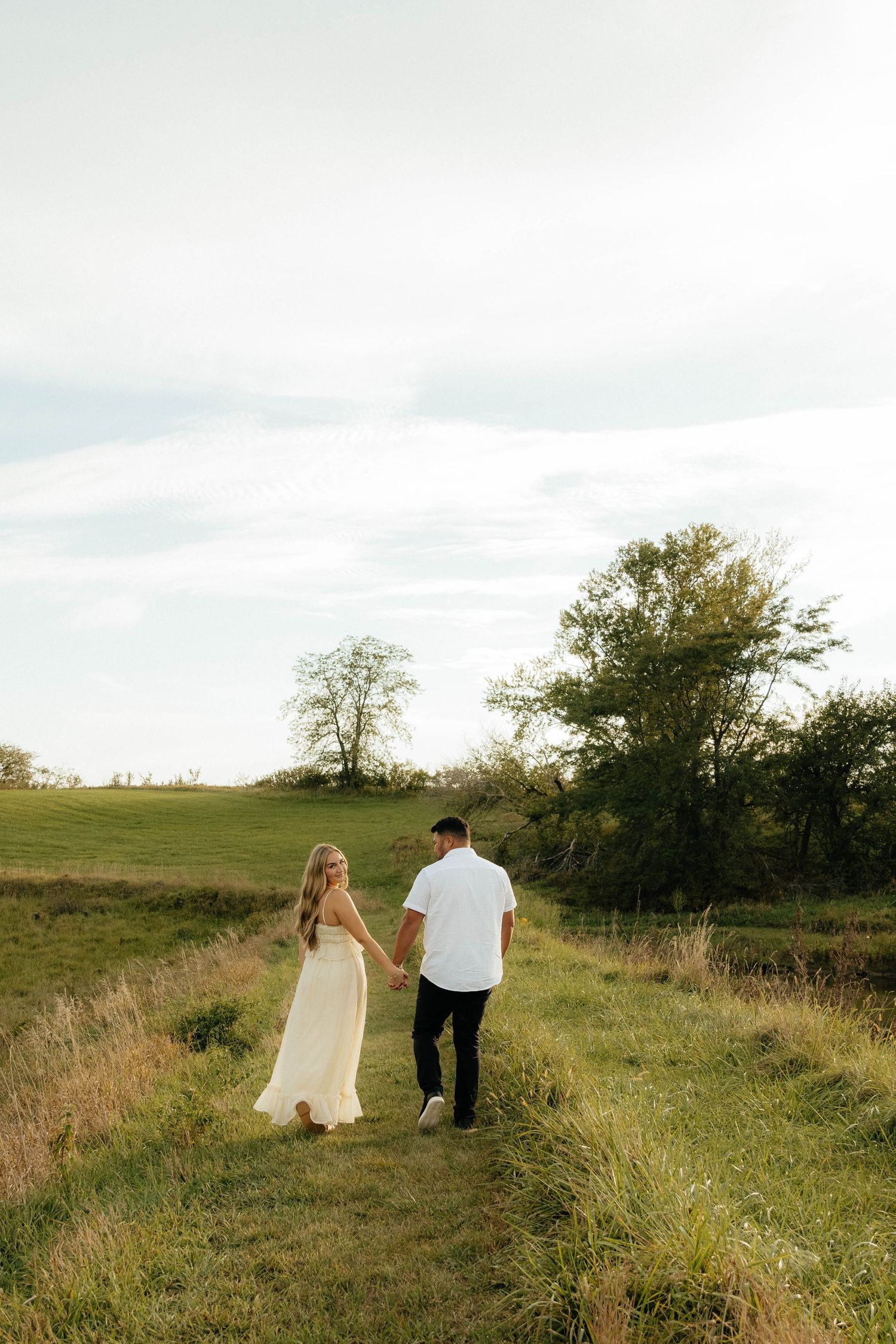 A couple taking engagement photos for their wedding in Iowa walks hand in hand through serene meadow under cloudy sky.