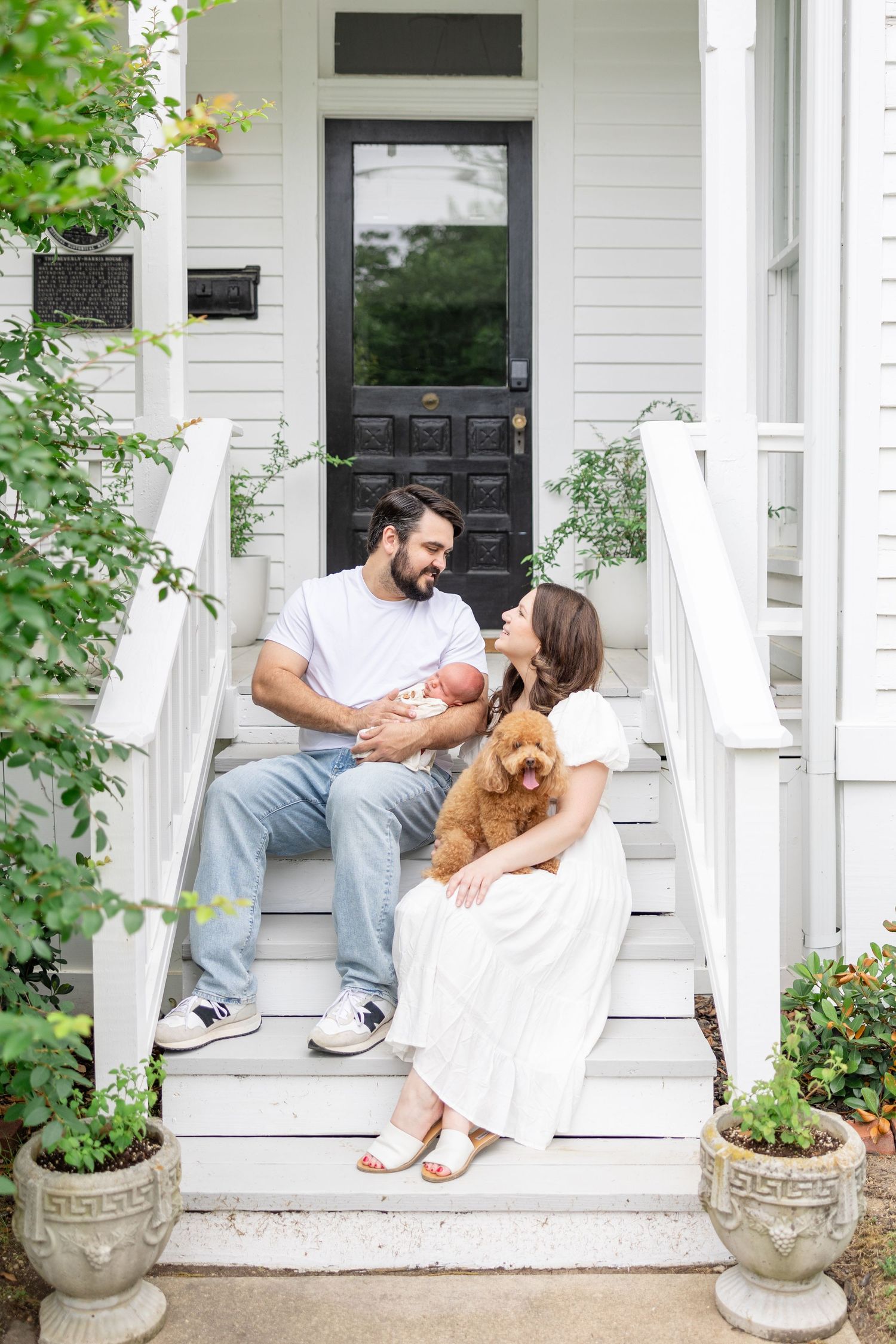 Happy parents in doorway holding newborn baby girl in pink while toddler runs toward them.