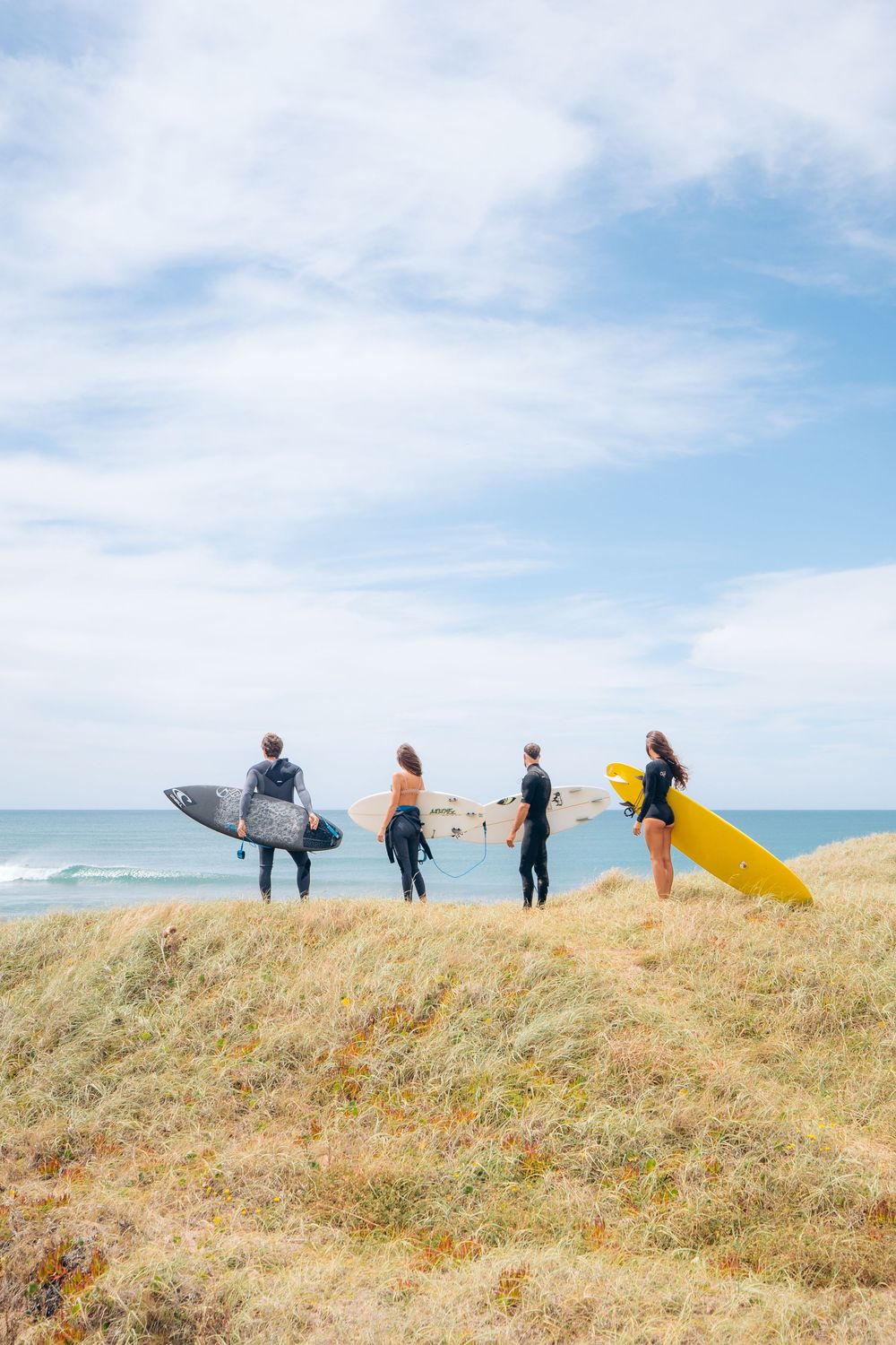 A group of surfers carrying surfboards walk along a grassy coastal hill overlooking the ocean.