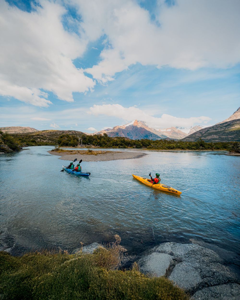 Two kayakers paddle on a scenic mountain lake surrounded by rugged peaks and blue skies.