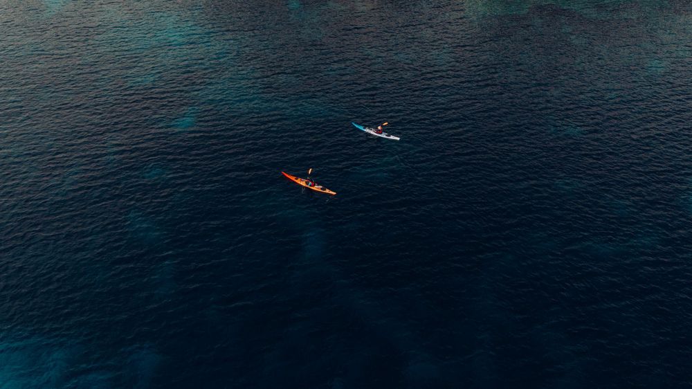 Aerial view of kayaks paddling on dark blue ocean waters surrounded by distant mountains.