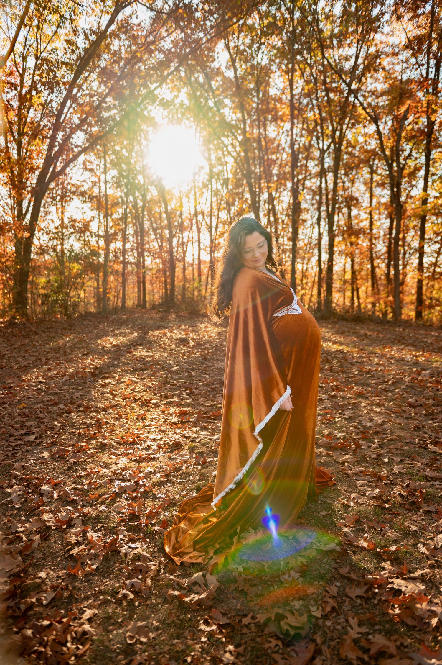 Maternity photo of expecting mother in flowing gown surrounded by golden autumn trees at sunset in the woods.