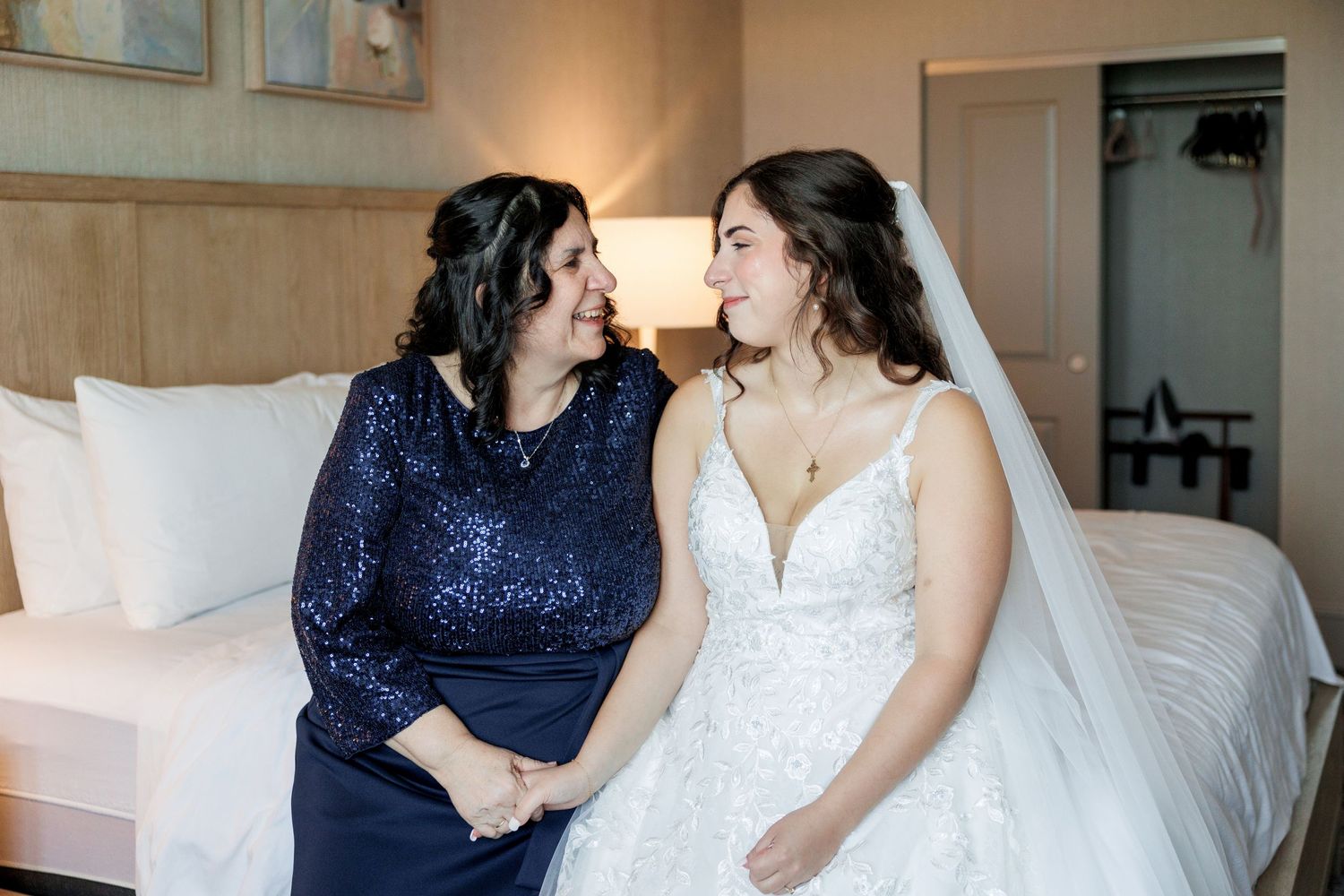 Bride in white gown and woman in navy dress share intimate moment while getting ready.