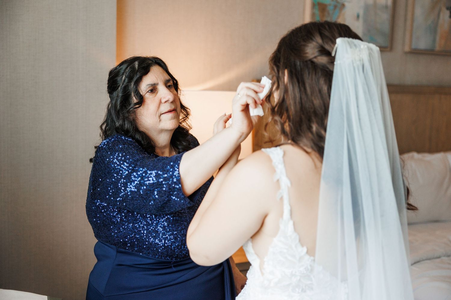 Woman in sparkling blue dress helps bride with final wedding preparations.