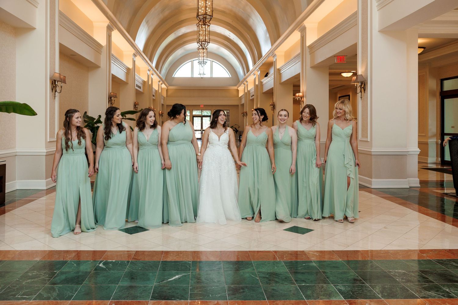 Bridal party in mint green dresses poses with bride in grand hallway with arched ceiling.