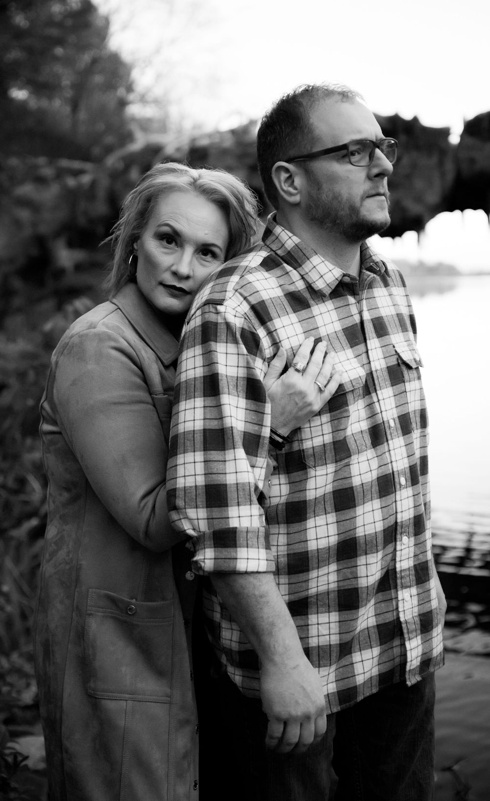 Black and white portrait of woman leaning on mans shoulder outdoors near water.