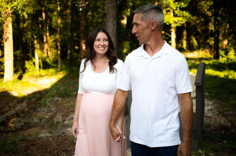 Pregnant woman in blush skirt holding hands with man in white shirt in sunny forest.
