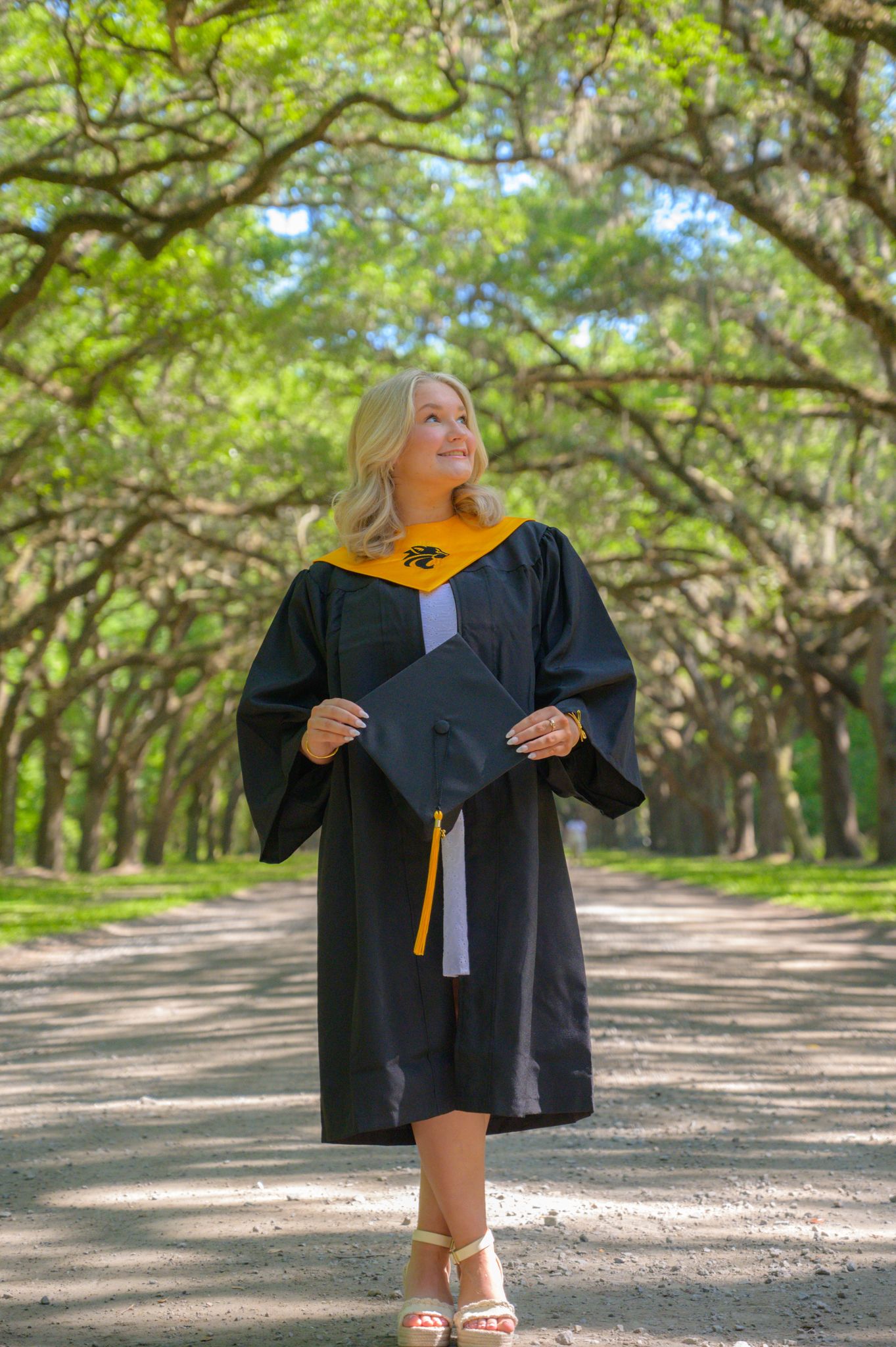 Graduate in cap and gown poses on tree-lined path during outdoor ceremony.