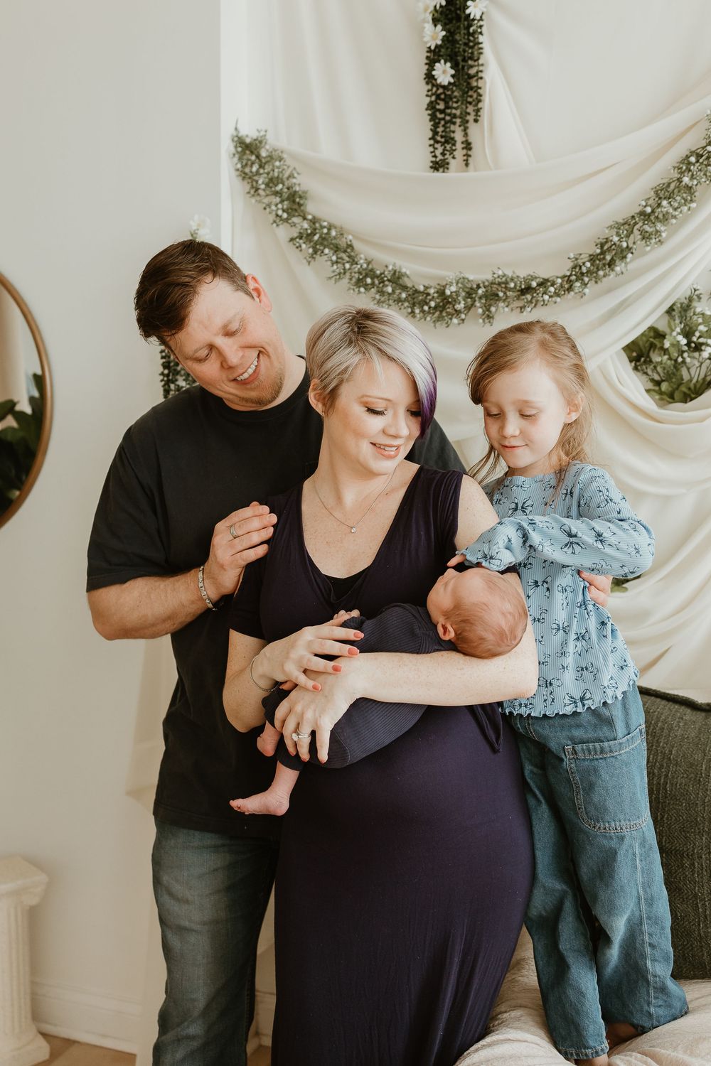 A family shares an intimate moment together in front of a white draped backdrop with greenery accents.