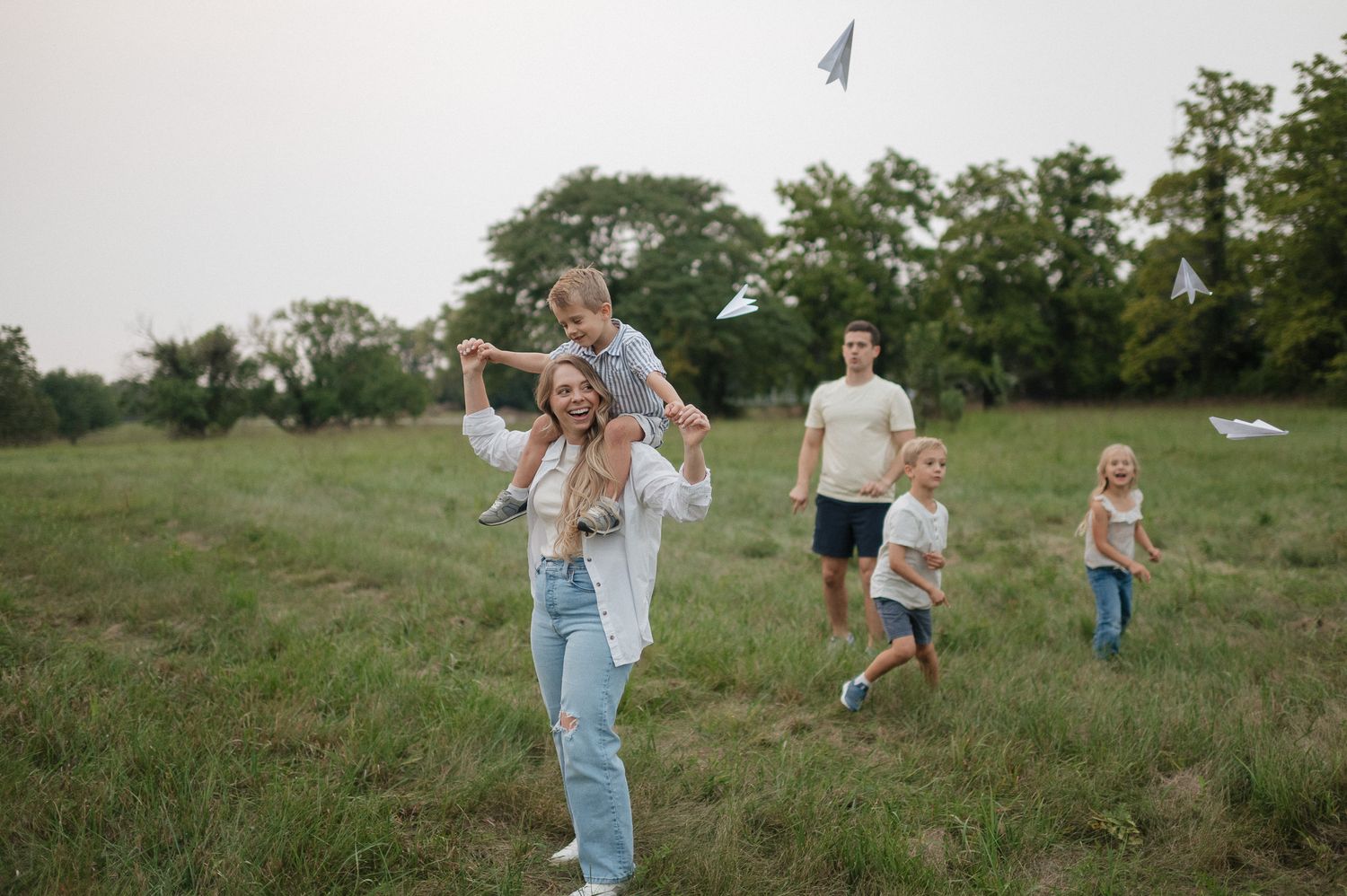 Kansas city family photographer giving her youngest son a shoulder ride while her other kids throw paper airplanes at shawnee mission park