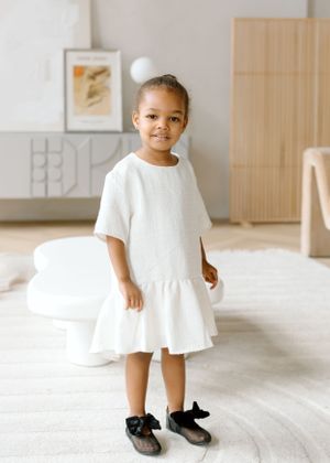 Young child stands in white dress and black shoes against minimalist home interior.