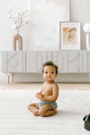 Baby in gray diaper sits on white carpet in front of modern decorative sideboard.