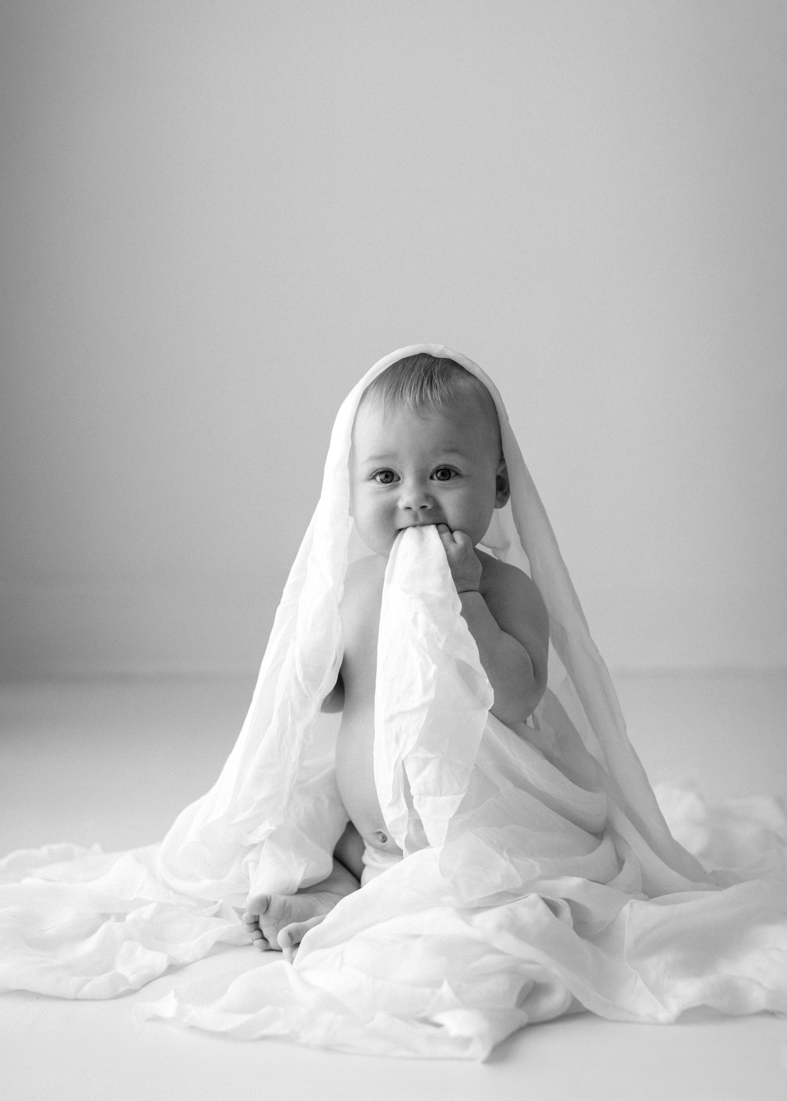 Black and white photo of a baby draped in a white blanket sitting on white fabric with a playful expression.