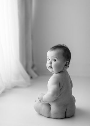 Black and white portrait of a chubby baby sitting on a bare floor near a window with soft natural lighting.