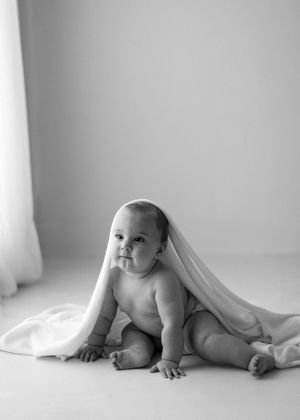 Black and white portrait of a baby sitting under a white blanket in a minimalist setting.