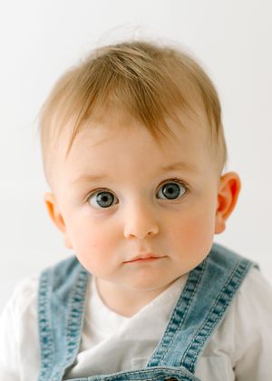 Baby in denim overalls with bright blue eyes gazes directly at camera.