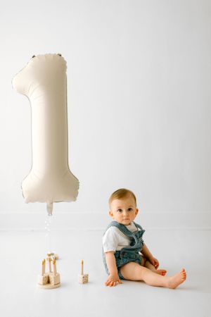 Baby in blue overalls sits beside large number one balloon against white backdrop.