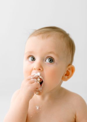 Baby with messy face enjoys eating cake while looking up with bright eyes.