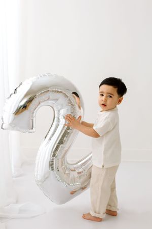 Young child holds large silver number 2 balloon in all-white setting.