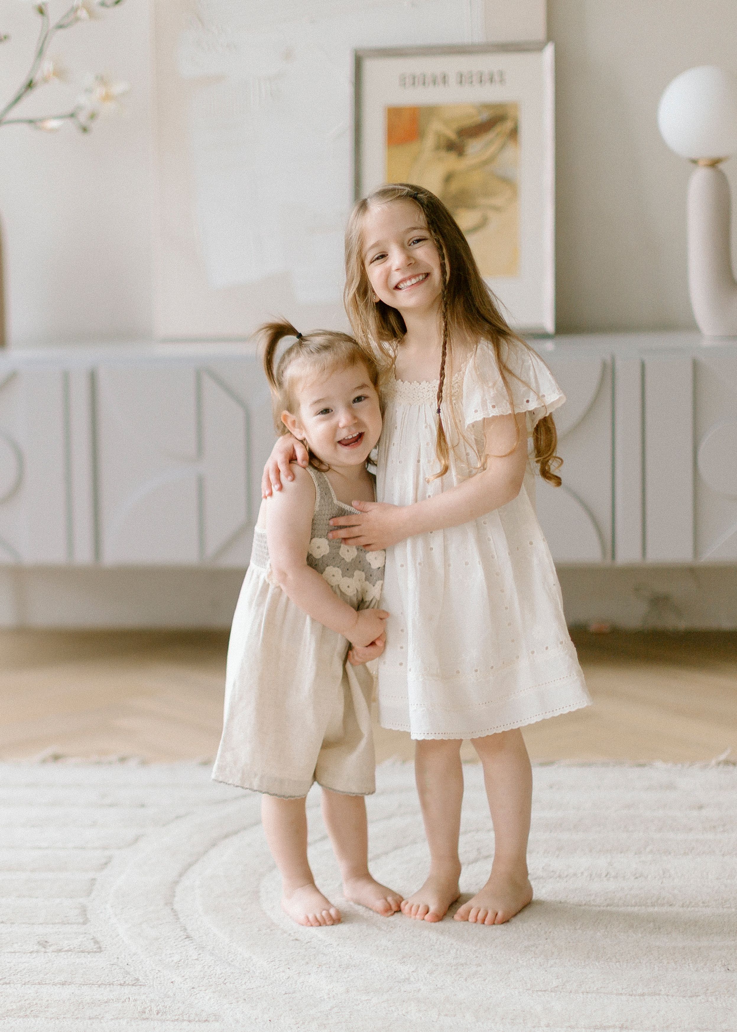 Two young children wearing white dresses stand barefoot together on a light carpet in front of wainscoting and artwork.