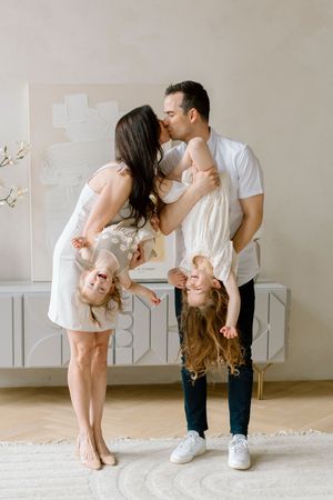 Playful family portrait with parents in white outfits holding two laughing children upside down in a minimalist room.