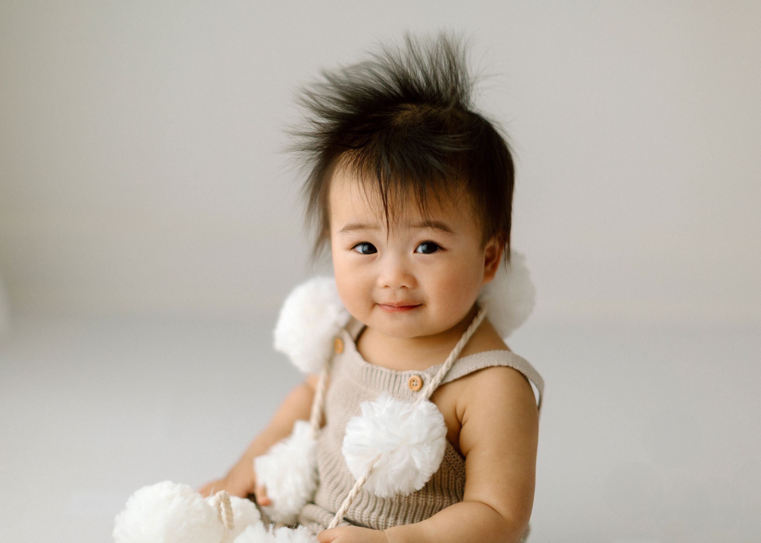 Smiling baby in beige knit tank top holds fluffy white toy against neutral background.