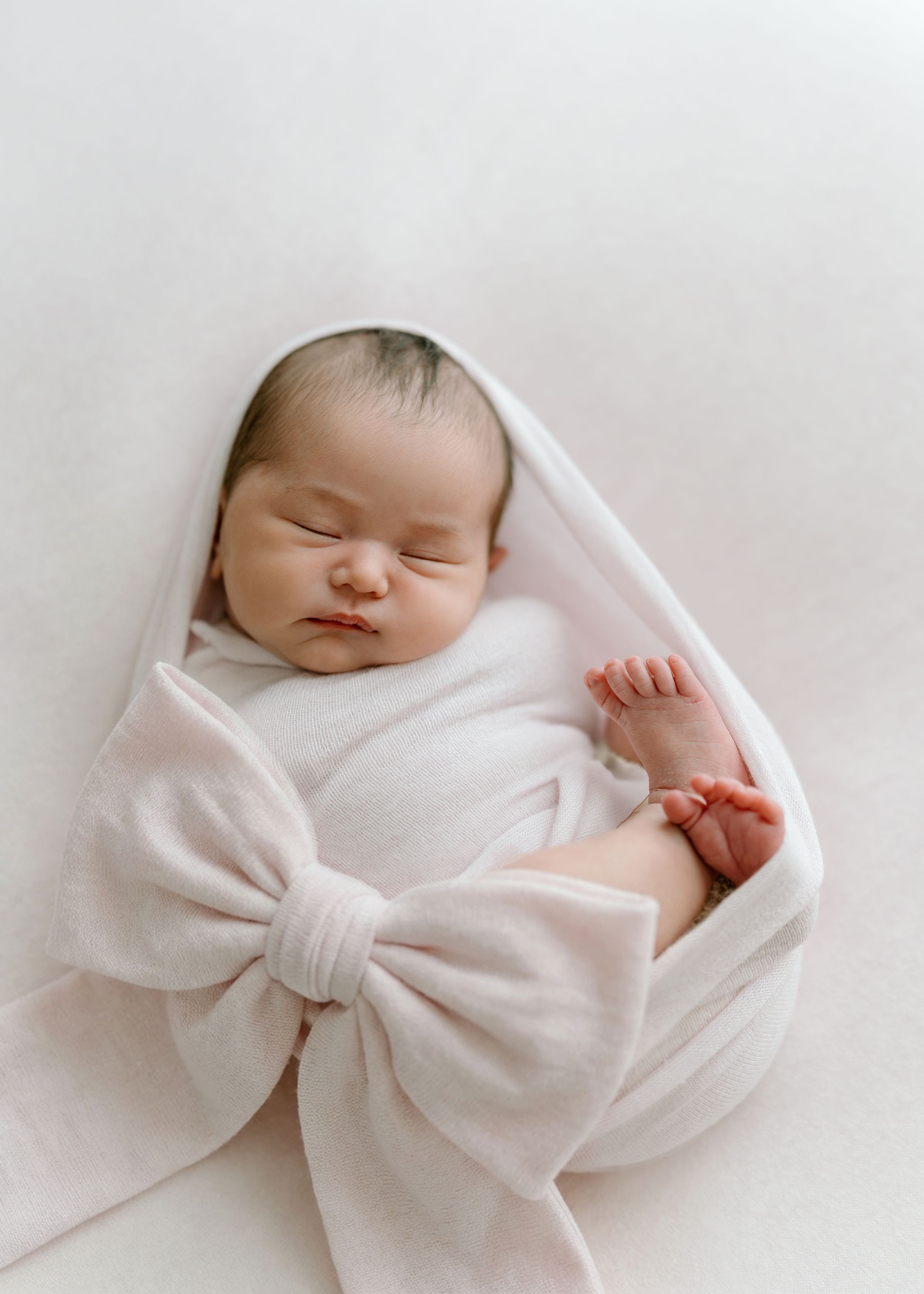 A sleeping newborn baby wrapped in white fabric with a large decorative bow headband.