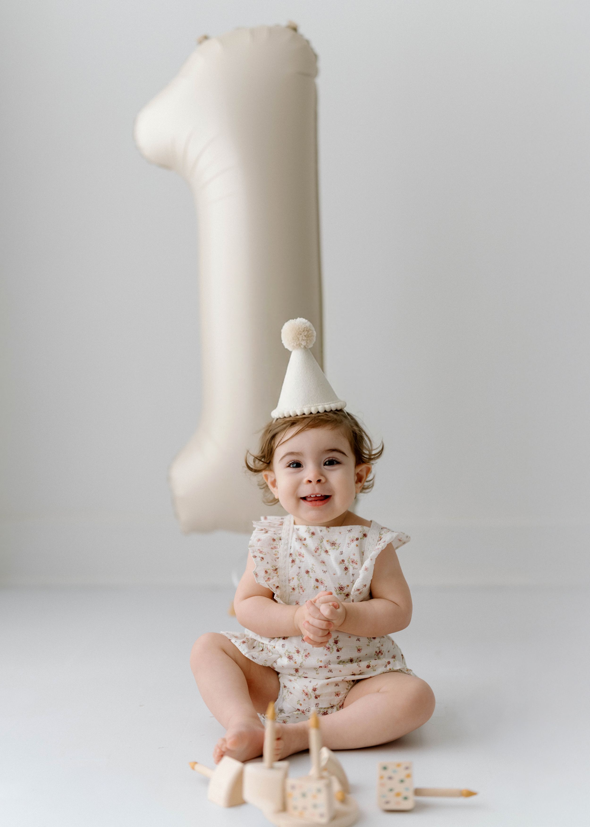 Baby wearing floral romper and party hat sits by large number one balloon with wooden toys on white background.