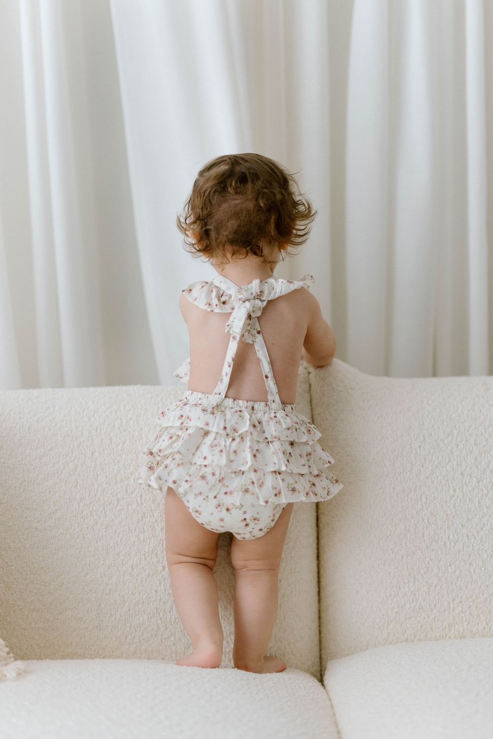 A toddler in a floral romper stands near a white curtain and sofa in a series of candid poses.