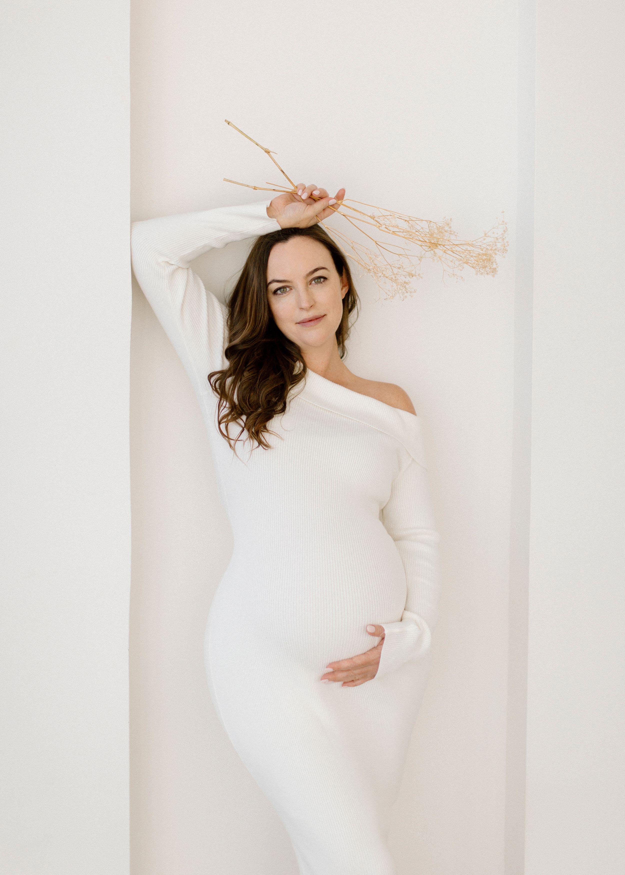A woman in an elegant white off-shoulder dress poses gracefully against a light-colored wall.