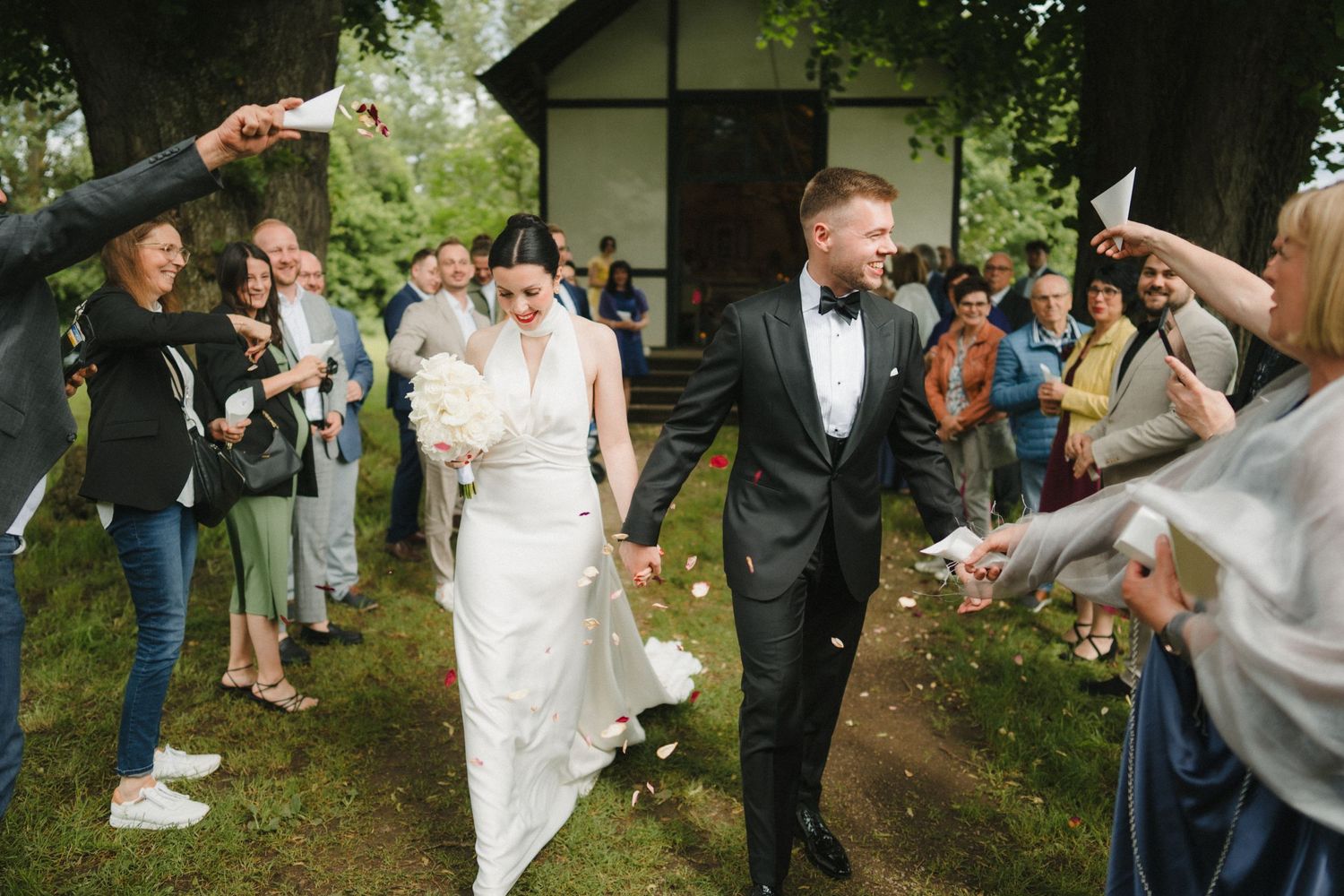 Wedding guests toss confetti over the couple walking down a garden path.
