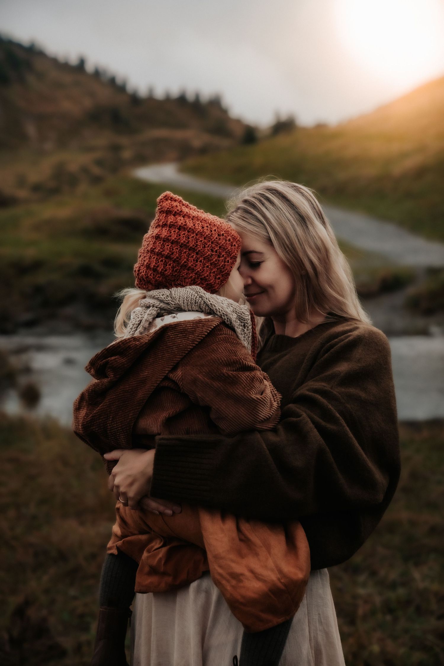 Parent and child share tender moment outdoors against mountain landscape at sunset.