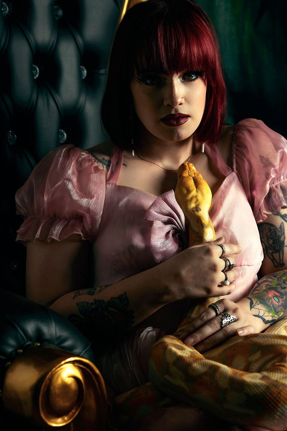 Tattooed woman with red hair in pink dress holding golden snake on dark tufted chair.